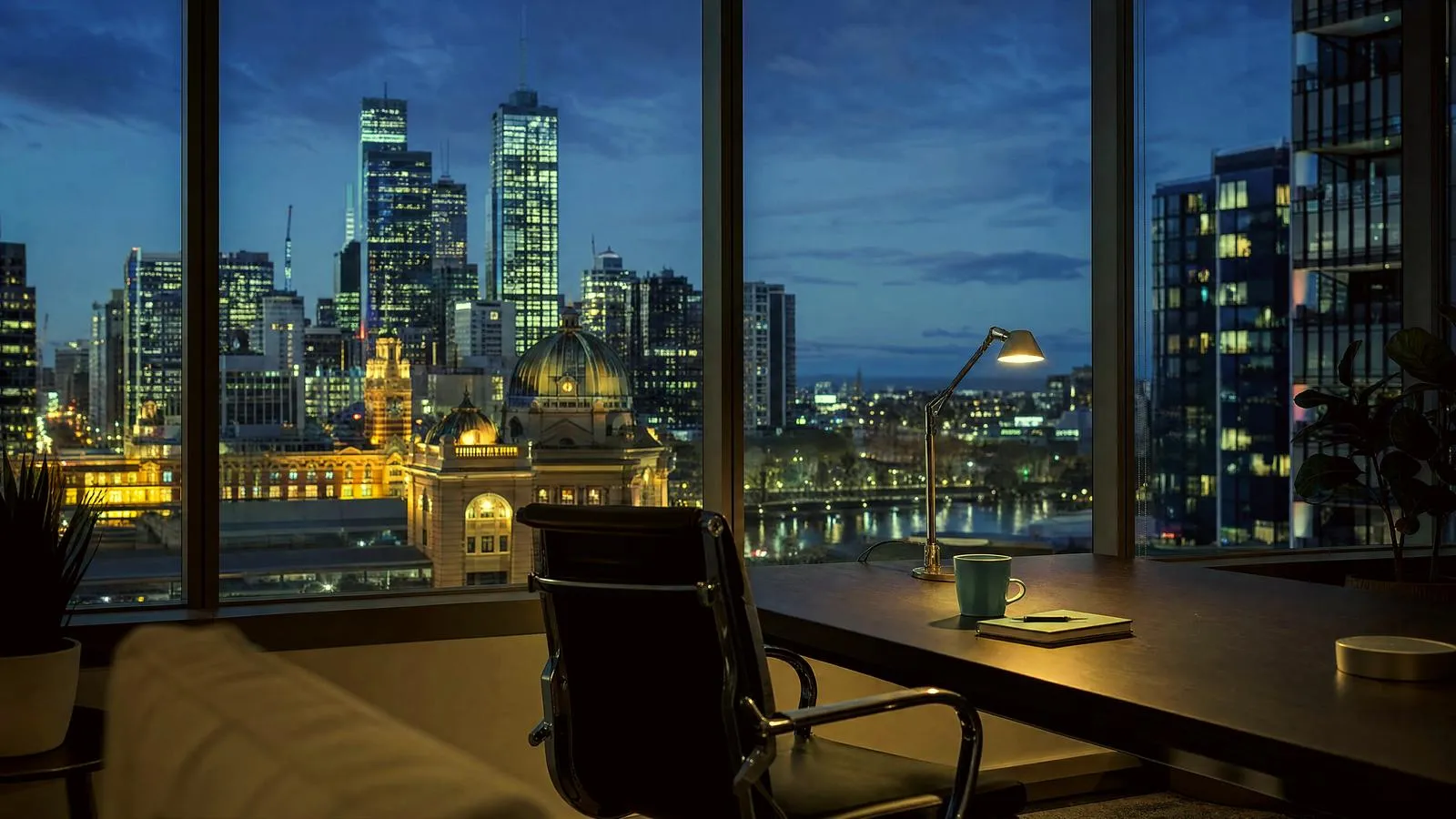 An empty Southbank office at dusk, a single desk lamp on and the lit Melbourne CBD and Yarra River beyond the windows.